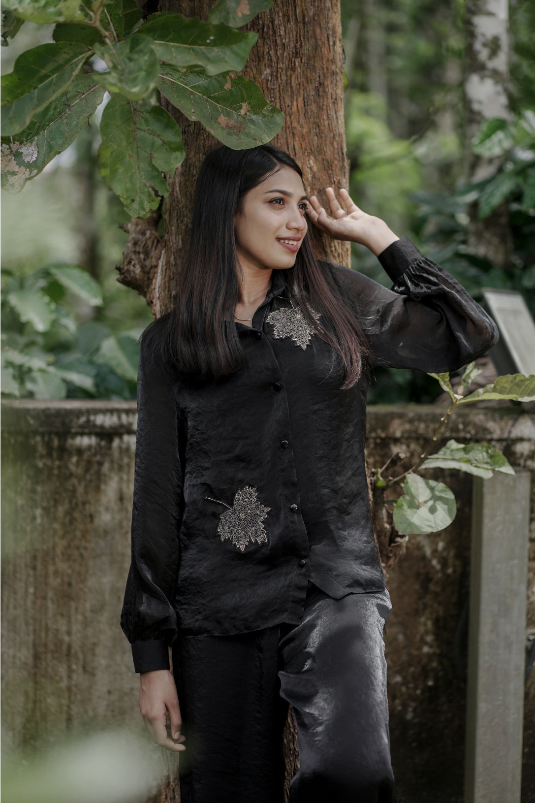 Woman wearing Noira black co-ord set with delicate leaf handwork embroidery, full balloon sleeves, standing near a tree