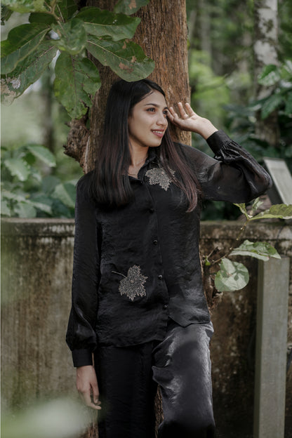 Woman wearing Noira black co-ord set with delicate leaf handwork embroidery, full balloon sleeves, standing near a tree