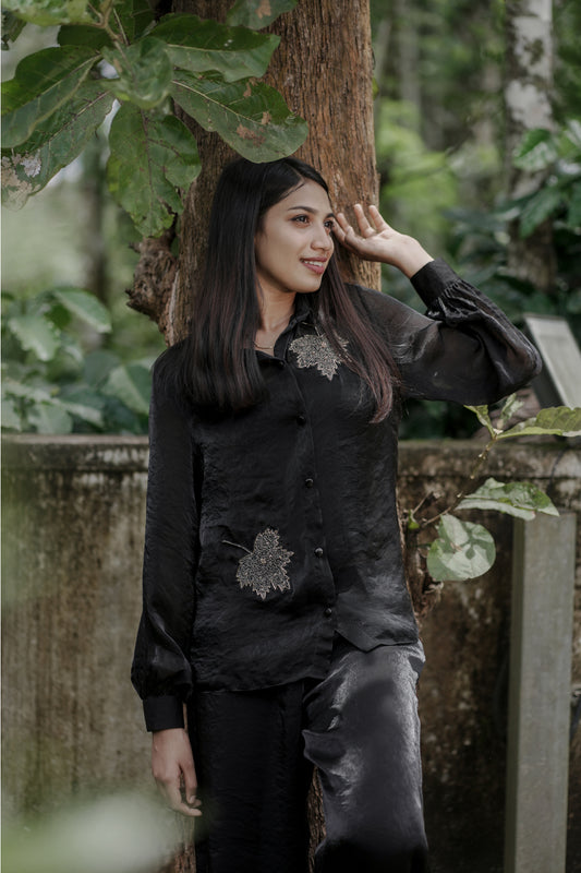 Woman wearing Noira black co-ord set with delicate leaf handwork embroidery, full balloon sleeves, standing near a tree