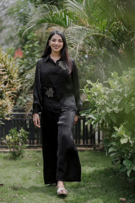 Woman wearing Noira black co-ord set with leaf handwork, full balloon sleeves and collar neck, standing in a garden with greenery