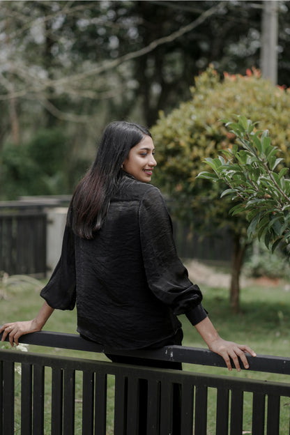 Woman wearing Noira black co-ord set, showing back view with full balloon sleeves, leaning on a fence with greenery in background