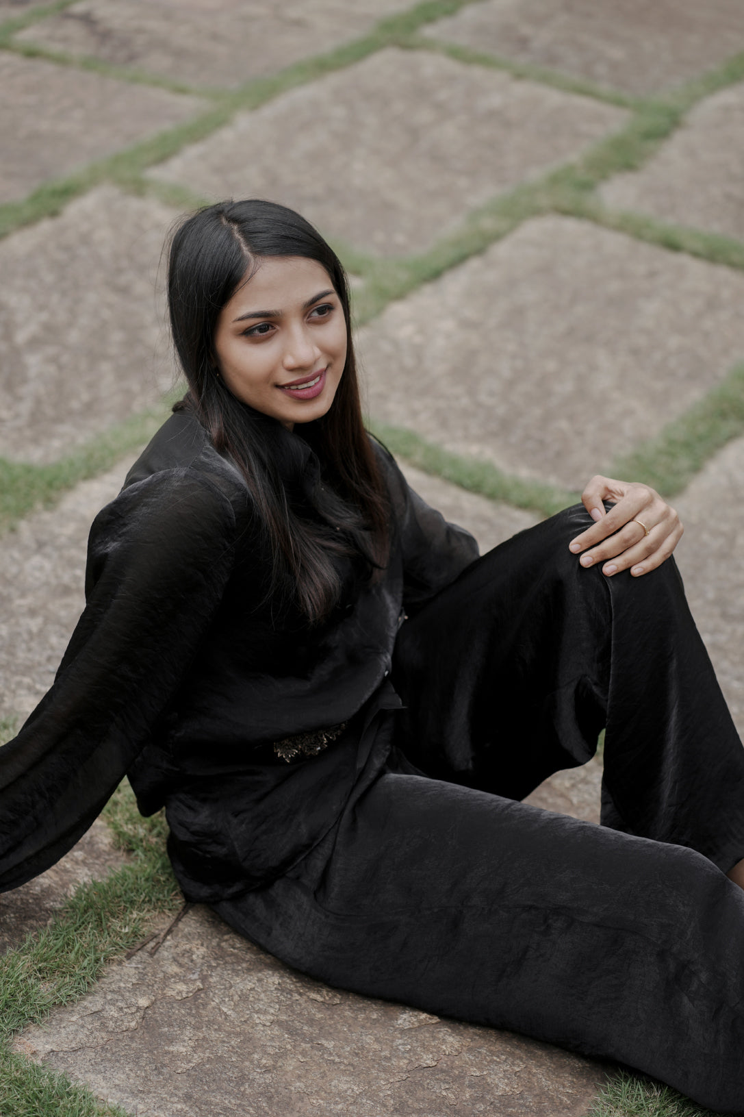 Woman wearing Noira black co-ord set sitting on stone pavement, showing flowy palazzo pants and balloon sleeves