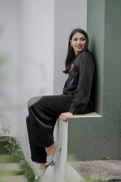 Woman wearing Noira black co-ord set sitting on a ledge against a plain wall, showing flowy palazzo pants and balloon sleeves