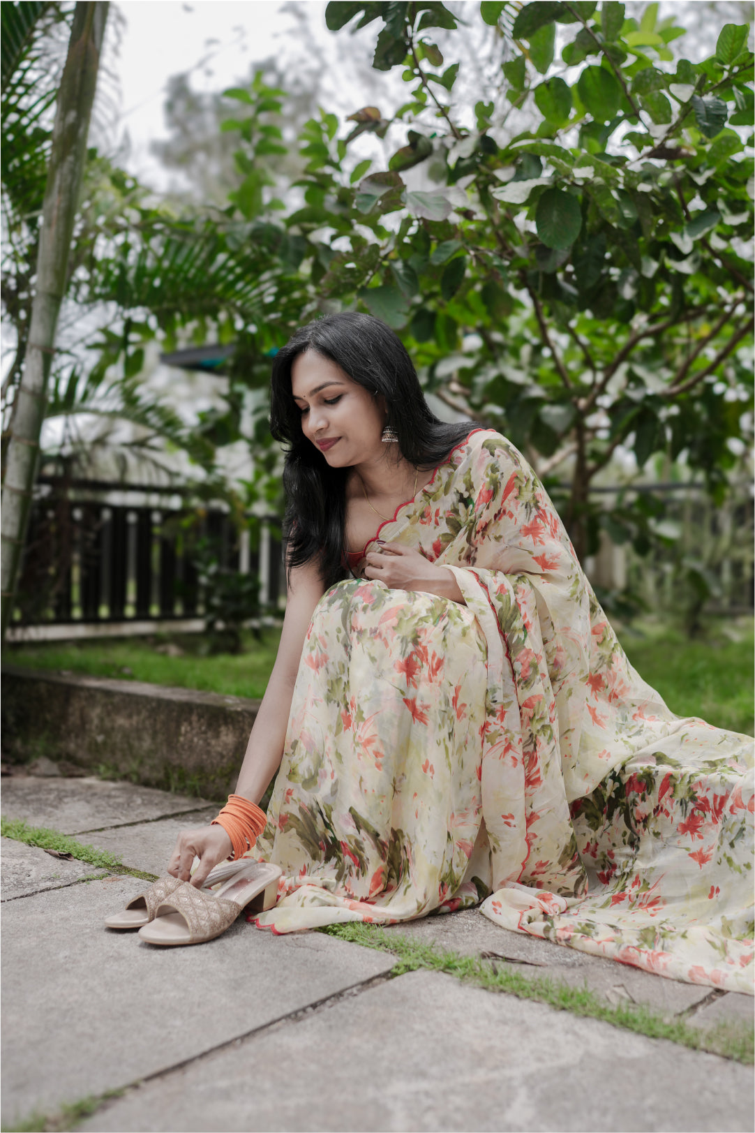 Woman sitting in Elaria saree with orange digital floral print on stone path with greenery backdrop