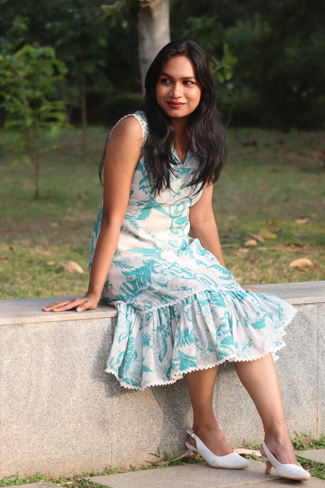Woman wearing Petal Aija Blue linen dress sitting on stone wall showing ruffled skirt and dress details