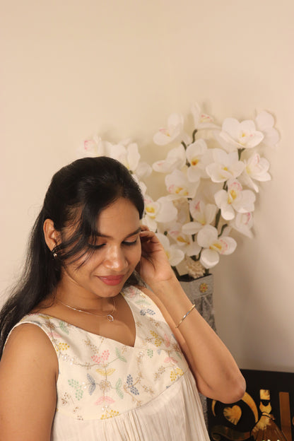 Woman wearing off-white sleeveless top with pastel embroidered yoke, looking down while adjusting her hair, white orchid flowers in background