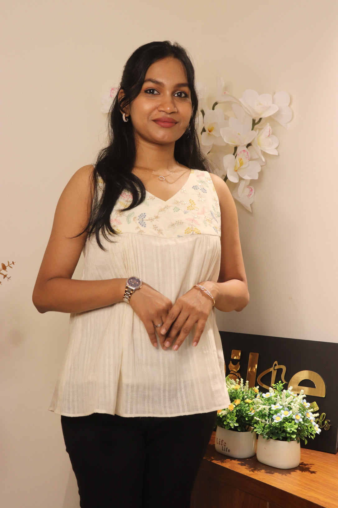 Woman wearing an off-white sleeveless top with pastel embroidered yoke and v-neck, hands folded at front